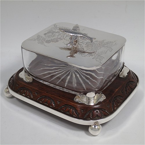 A very handsome Antique Victorian Silver Plated sardine dish, having a rectangular body with rounded corners, a cut-crystal liner sitting in a frame attached to an Oak wooden base with four ball feet, a lift-off lid with cast crossed sardines above hand-engraved floral decoration. Made in ca. 1890. The dimensions of this fine hand-made antique silver-plated sardine dish are length 18 cms (7 inches), width 15 cms (6 inches), and height 10 cms (4 inches).   