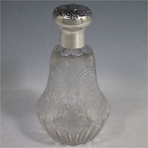 An Antique Victorian Sterling Silver and hand-cut crystal large table scent bottle, having a hand-chased round hinged lid with floral and figural decoration, a plain round mount, an internal stopper, and an unusual shaped hand-cut crystal body with floral, swags, bows, and fluted decoration. Made by William Comyns of London in 1900. The dimensions of this fine hand-made antique silver & crystal scent bottle are height 18 cms (7 inches), and diameter 10 cms (4 inches).    