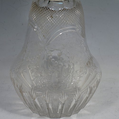 An Antique Victorian Sterling Silver and hand-cut crystal large table scent bottle, having a hand-chased round hinged lid with floral and figural decoration, a plain round mount, an internal stopper, and an unusual shaped hand-cut crystal body with floral, swags, bows, and fluted decoration. Made by William Comyns of London in 1900. The dimensions of this fine hand-made antique silver & crystal scent bottle are height 18 cms (7 inches), and diameter 10 cms (4 inches).    