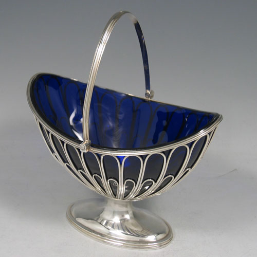 Antique Georgian sterling silver sugar basket, having an oval wire-work body, with hinged reeded swing-handle, a blue-glass liner, and sitting on a pedestal foot. Made by Robert Hennell I of London in 1792. The dimensions of this fine hand-made silver sugar basket are height (inc. handle) 18 cms (7 inches), length 15.5 cms (6 inches), and it weighs approx. 222g (7.2 troy ounces).