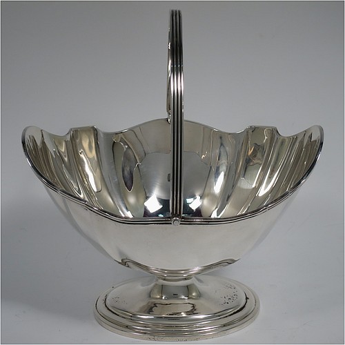 An Antique Sterling Silver sugar basket in a Georgian style, having a shaped oval body with panelled shoulders, an applied reeded border, a reeded swing-handle, and sitting on a pedestal foot. Made by Thomas Bradbury and Sons of Sheffield in 1913. The dimensions of this fine hand-made antique silver sugar basket are height 17 cms (6.75 inches), length 15 cms (6 inches), width 12.5 cms (5 inches), and it weighs approx. 231g (7.5 troy ounces).    