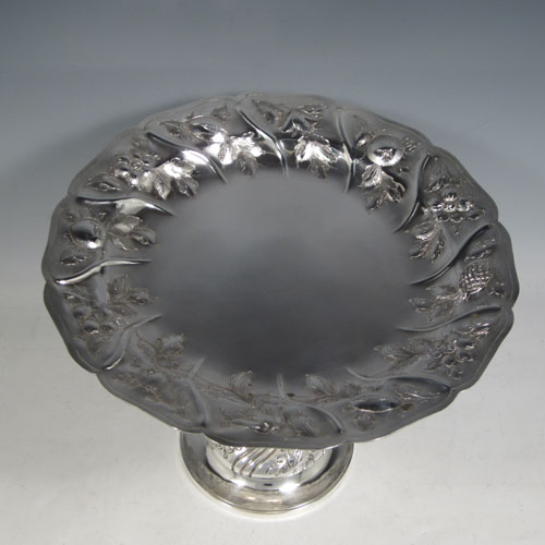 Antique Victorian sterling silver tazza dish, having a round body with hand-chased floral and fruit decoration, sitting on a pedestal foot with matching decoration and swirl fluting. Made by Henry Holland of London in 1874. The dimensions of this fine hand-made silver tazza dish are height 21 cms (8.25 inches), diameter 26 cms (10.25 inches), and it weighs approx. 838g (27 troy ounces).