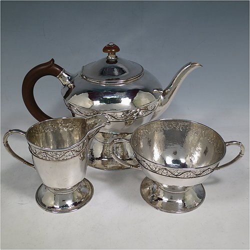 A unique Sterling Silver Arts and Crafts style three-piece tea set, having round bodies with hand-hammered decoration, with applied and hand-chased bands of floral work, the teapot with brown-stained wooden handle and finial, and the sugar and creamer with cast rounded handles, all with applied rope-twist borders, and all sitting on spreading pedestal feet. This beautiful silver tea set was made by Harold Edwin Landon of Chester in 1933. The dimensions of this fine hand-made Arts and Crafts tea service are height of teapot 18 cms (7 inches), length 28 cms (11 inches), diameter 15 cms (6 inches), and with a total weight of approx. 992g (32 troy ounces).