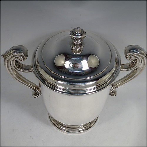 A very handsome and heavy Antique Victorian Sterling Silver trophy cup and cover, in a George II style, having a plain round body, two flying scroll side-handles with anthemion leaf thumb-pieces, a pull-off lid with cast finial, and all sitting on a stepped pedestal foot. Made by William Barnard of London in 1895. The dimensions of this fine hand-made antique silver trophy cup and cover are height 27 cms (10.5 inches), spread across arms 30.5 cms (12 inches), and it weighs approx. 1,787g (57.6 troy ounces).   