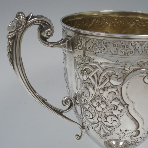 A very beautiful Antique Victorian Sterling Silver trophy cup, having a round body with tapering sides all hand-chased with floral and scroll decoration surrounding vacant cartouches either side, two cast scroll handles with figural thumb-pieces, a gold-gilt interior, and all sitting on a pedestal foot. Made by John Edward Bingham and Charles Henry Bingham of Sheffield in 1876. The dimensions of this fine hand-made antique silver trophy cup are height 24 cms (9.5 inches), spread across arms 22 cms (8.75 inches), and it weighs approx. 620g (20 troy ounces).   