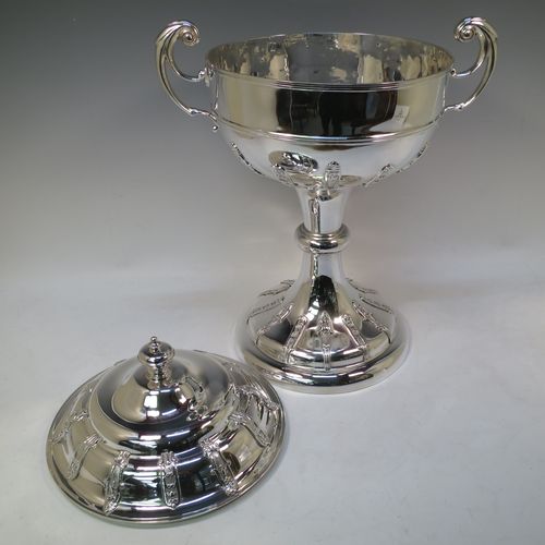 A very impressive and large Antique Edwardian  Sterling Silver trophy cup and cover, having a round body with applied floral strap-work decoration, two flying scroll side-handles, a central applied girdle band, a pull-off lid with matching applied strap-work and a cast round finial, and all sitting on a stepped pedestal foot, again, with matching decoration. Made by Alexander Clark of Sheffield in 1906. The dimensions of this fine hand-made antique silver trophy cup and cover are height 48 cms (19 inches), spread across arms 37 cms (14.5 inches), and it weighs approx. 2,842g (92 troy ounces). Please note that this item has a full armorial crest engraved on one side. 