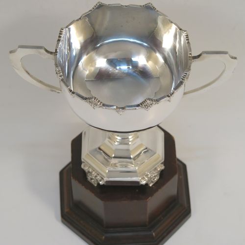 A small but very handsome Sterling Silver trophy cup, having a plain round bellied body, with two scroll side-handles, an applied cast floral border, a panelled pedestal foot with four cast floral legs, and with its original brown wooden panelled plinth. This elegant silver trophy cup and plinth was made by the E. E. Poston and Co.,  of Birmingham in 1935. The dimensions of this fine hand-made silver trophy cup are height (without plinth) 12 cms (4.75 inches), spread across arms 14.5 cms (5.75 inches), and it weighs approx. 171g (5.5 troy ounces).  