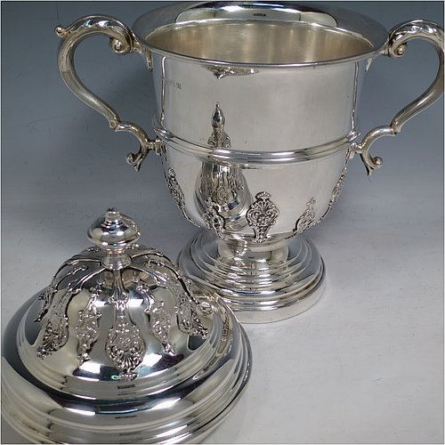 A Sterling Silver trophy cup and cover, in a George II style, having a round body with applied floral strap-work decoration, two scroll side-handles with anthemion leaf thumb-pieces, a central applied girdle band, a pull-off lid with matching applied strap-work and a cast round finial, and all sitting on a stepped pedestal foot. Made by George Bryan & Co., of Birmingham in 1936. The dimensions of this fine hand-made silver trophy cup and cover are height 30 cms (11.75 inches), spread across arms 26.5 cms (10.5 inches), and it weighs approx. 1,132g (36.5 troy ounces).    