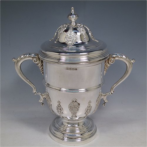 A Sterling Silver trophy cup and cover, in a George II style, having a round body with applied floral strap-work decoration, two scroll side-handles with anthemion leaf thumb-pieces, a central applied girdle band, a pull-off lid with matching applied strap-work and a cast round finial, and all sitting on a stepped pedestal foot. Made by J. A. Restall of Birmingham in 1926. The dimensions of this fine hand-made silver trophy cup and cover are height 26 cms (10.25 inches), spread across arms 24 cms (9.5 inches), and it weighs approx. 830g (27 troy ounces).    