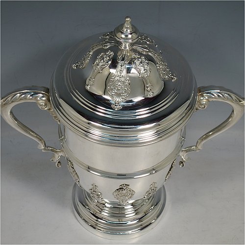 A Sterling Silver trophy cup and cover, in a George II style, having a round body with applied floral strap-work decoration, two scroll side-handles with anthemion leaf thumb-pieces, a central applied girdle band, a pull-off lid with matching applied strap-work and a cast round finial, and all sitting on a stepped pedestal foot. Made by J. A. Restall of Birmingham in 1926. The dimensions of this fine hand-made silver trophy cup and cover are height 26 cms (10.25 inches), spread across arms 24 cms (9.5 inches), and it weighs approx. 830g (27 troy ounces).    