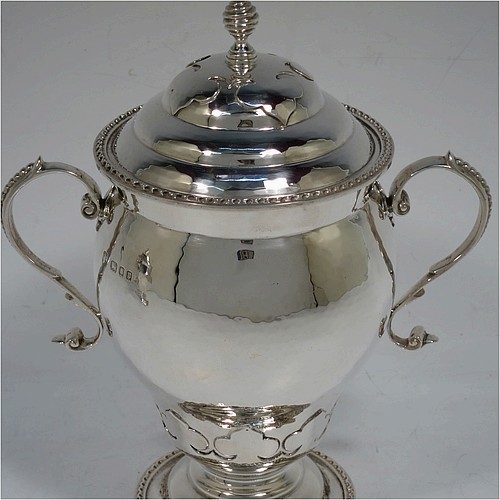 A very unusual pair of Sterling Silver trophy cups and covers, in an Arts and Crafts style, having round bellied bodies with hand-hammered decoration, applied zigzag borders and cut-card work, two side scroll handles with beaded thumb-pieces, removable covers with spiral finials, and all sitting on stepped pedestal feet. Made by D. and J. Wellby of London in 1931. The dimensions of these fine hand-made silver trophy cups and covers are height 17.5 cms (7 inches), spread across arms 15 cms (6 inches), and they weigh a total of  approx. 825g (26.6 troy ounces).   