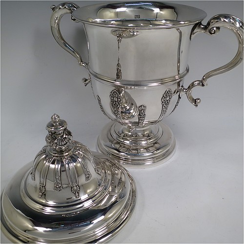 A very handsome Antique Edwardian Sterling Silver trophy cup and cover, in a George II style, having a round body with applied floral strap-work decoration, two scroll side-handles with anthemion leaf thumb-pieces, a central applied girdle band, a pull-off lid with matching applied strap-work and a cast round floral finial, and all sitting on a stepped pedestal foot. Made by Walker and Hall of Sheffield in 1909. The dimensions of this fine hand-made antique silver trophy cup and cover are height 37 cms (14.5 inches), spread across arms 33 cms (13 inches), and it weighs approx. 1,570g (51 troy ounces).    