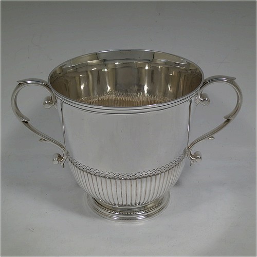 A very handsome Sterling Silver trophy cup in a George III neoclassical style, having a round body with tapering sides and half-fluted decoration, with hand-chased reeded borders, two cast scroll side-handles, and all sitting on a pedestal foot. Made by Charles and Richard Comyns of London in 1921. The dimensions of this fine hand-made silver trophy cup are height 14.5 cms (5.75 inches), spread across arms 24 cms (9.5 inches), and it weighs approx. 683g (22 troy ounces).    