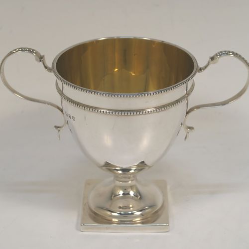 A small but very elegant Antique Victorian Sterling Silver trophy cup in a George III neoclassical style, having a round plain body with tapering sides, a gold-gilt interior, with two applied bead top borders, two scroll side-handles,  and all sitting on a pedestal foot with a square base. This handsome antique silver trophy cup was made by Angel and Savory of London in 1890. The dimensions of this fine hand-made antique silver trophy cup are height 12 cms (4.75 inches), spread across arms 16 cms (6.3 inches), and it weighs approx. 182g (5.9 troy ounces).  