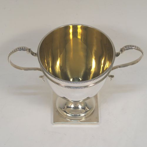 A small but very elegant Antique Victorian Sterling Silver trophy cup in a George III neoclassical style, having a round plain body with tapering sides, a gold-gilt interior, with two applied bead top borders, two scroll side-handles,  and all sitting on a pedestal foot with a square base. This handsome antique silver trophy cup was made by Angel and Savory of London in 1890. The dimensions of this fine hand-made antique silver trophy cup are height 12 cms (4.75 inches), spread across arms 16 cms (6.3 inches), and it weighs approx. 182g (5.9 troy ounces).  