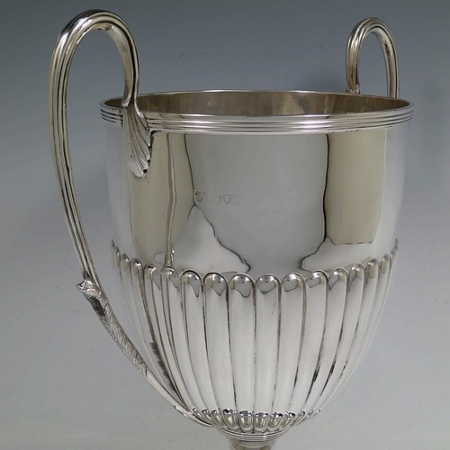 A very handsome Antique Victorian Sterling Silver trophy cup in a George III neoclassical style, having a round body with tapering sides and half-fluted decoration, with hand-chased reeded borders, two looped side-handles with anthemion leaves, and all sitting on a pedestal foot. Made by Charles Stuart Harris of London in 1895. The dimensions of this fine hand-made antique silver trophy cup are height 32.5 cms (12.75 inches), spread across arms 26.5 cms (10.5 inches), and it weighs approx. 940g (30 troy ounces).    