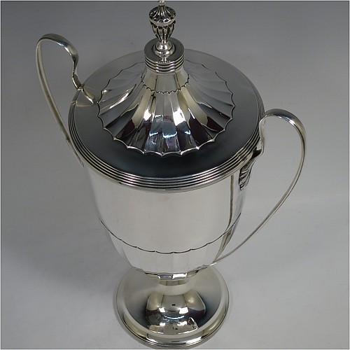 A very handsome Sterling Silver trophy cup and cover in a George III neoclassical style, having a round body with tapering sides and half-fluted decoration, with hand-chased reeded borders, two looped side-handles, an original cover with matching decoration and cast urn finial, and all sitting on a pedestal foot. Made by Robert Pringle  of London in 1923. The dimensions of this fine hand-made silver trophy cup and cover are height 33 cms (13 inches), spread across arms 21 cms (8.25 inches), and it weighs approx. 879g (28 troy ounces).    