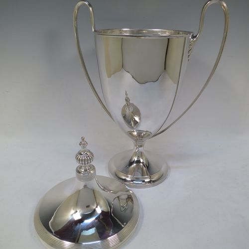 A very handsome and large Sterling Silver trophy cup and cover in a George III neocclassical style, having a round plain body with tapering sides, with hand-chased reeded borders, two looped side-handles with anthemion leaf ends, an original cover with matching decoration and cast knop finial, and all sitting on a pedestal foot. Made by James Dixon and Sons of Sheffield in 1926. The dimensions of this fine hand-made silver trophy cup and cover are height 42 cms (16.5 inches), spread across arms 28 cms (11 inches), and it weighs approx. 1,413g (45.6 troy ounces).    