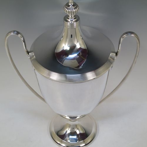 A very handsome and large Sterling Silver trophy cup and cover in a George III neocclassical style, having a round plain body with tapering sides, with hand-chased reeded borders, two looped side-handles with anthemion leaf ends, an original cover with matching decoration and cast knop finial, and all sitting on a pedestal foot. Made by James Dixon and Sons of Sheffield in 1926. The dimensions of this fine hand-made silver trophy cup and cover are height 42 cms (16.5 inches), spread across arms 28 cms (11 inches), and it weighs approx. 1,413g (45.6 troy ounces).    