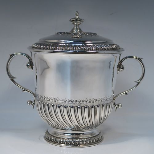 A very handsome Antique Edwardian Sterling Silver trophy cup and cover, in the shape of a lidded Porringer, having a round body with hand-chased half-fluted decoration, with two cast scroll side-handles, a lift-off cover with matching decoration and urn finial, and all sitting on a pedestal foot. This elegant silver cup and cover was made by Robert Comyns of London in 1910. The dimensions of this fine hand-made antique silver trophy porringer cup are height 21 cms (8.25 inches), spread across handles 24 cms (9.5 inches), and it weighs approx. 1,118g (36 troy ounces).   