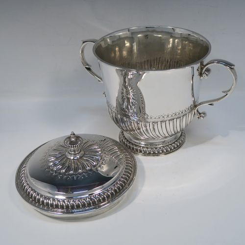 A very handsome Antique Edwardian Sterling Silver trophy cup and cover, in the shape of a lidded Porringer, having a round body with hand-chased half-fluted decoration, with two cast scroll side-handles, a lift-off cover with matching decoration and urn finial, and all sitting on a pedestal foot. This elegant silver cup and cover was made by Robert Comyns of London in 1910. The dimensions of this fine hand-made antique silver trophy porringer cup are height 21 cms (8.25 inches), spread across handles 24 cms (9.5 inches), and it weighs approx. 1,118g (36 troy ounces).   