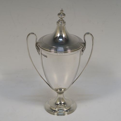 A very handsome Sterling Silver trophy cup and cover in a George III neoclassical style, having a round plain body with tapering sides, with hand-chased reeded borders, two looped side-handles, an original cover with matching decoration and cast knop finial, and all sitting on a pedestal foot. This beautiful silver trophy cup was made by Walker and Hall of Sheffield in 1927. The dimensions of this fine hand-made silver trophy cup and cover are height 18 cms (7 inches), spread across arms 11 cms (4.25 inches), and it weighs approx. 223g (7.2 troy ounces).   