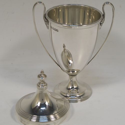 A very handsome Sterling Silver trophy cup and cover in a George III neoclassical style, having a round plain body with tapering sides, with hand-chased reeded borders, two looped side-handles, an original cover with matching decoration and cast knop finial, and all sitting on a pedestal foot. This beautiful silver trophy cup was made by Walker and Hall of Sheffield in 1927. The dimensions of this fine hand-made silver trophy cup and cover are height 18 cms (7 inches), spread across arms 11 cms (4.25 inches), and it weighs approx. 223g (7.2 troy ounces).   