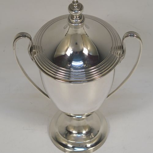 A very handsome Sterling Silver trophy cup and cover in a George III neoclassical style, having a round plain body with tapering sides, with hand-chased reeded borders, two looped side-handles, an original cover with matching decoration and cast knop finial, and all sitting on a pedestal foot. This beautiful silver trophy cup was made by Walker and Hall of Sheffield in 1927. The dimensions of this fine hand-made silver trophy cup and cover are height 18 cms (7 inches), spread across arms 11 cms (4.25 inches), and it weighs approx. 223g (7.2 troy ounces).   