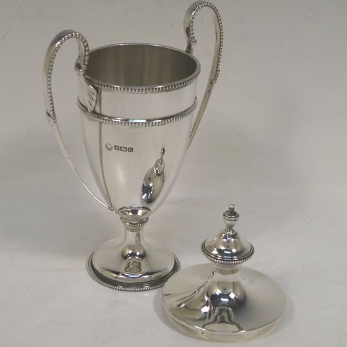 A small but very elegant Antique Sterling Silver trophy cup and cover in a George III neoclassical style, having a round plain body with tapering sides, with applied bead-edged borders, two looped side-handles, an original cover with matching decoration and cast urn finial, and all sitting on a pedestal foot. This handsome silver trophy cup was made by William Hutton of Sheffield in 1912. The dimensions of this fine hand-made silver trophy cup and cover are height 20 cms (8 inches), spread across arms 12.5 cms (5 inches), and it weighs approx. 280g (9 troy ounces).   