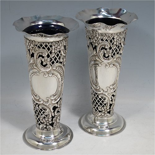 An Antique Victorian Sterling Silver pair of vases, having hand-chased and pierced bodies with floral decoration on round tapering sides, with vacant cartouches on one side, removable blue-glass liners, with wavy-edge rims and and sitting on pedestal feet. Made by the William Comyns of London in 1897. The dimensions of this fine hand-made pair of antique silver vases are height 18 cms (7 inches), diameter at top 9 cms (3.5 inches), and they weigh a total of 272g (8.8 troy ounces).   