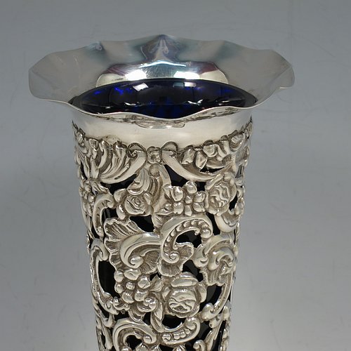 An Antique Victorian Sterling Silver pair of vases, having hand-chased and pierced bodies with floral decoration on round tapering sides, with vacant cartouches on one side, removable blue-glass liners, with wavy-edge rims and and sitting on pedestal feet. Made by the William Comyns of London in 1897. The dimensions of this fine hand-made pair of antique silver vases are height 18 cms (7 inches), diameter at top 9 cms (3.5 inches), and they weigh a total of 272g (8.8 troy ounces).   