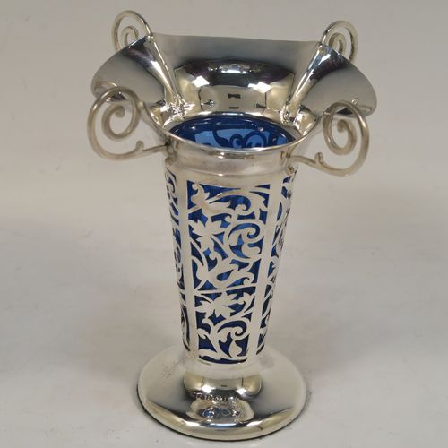 A vert pretty pair of Antique Edwardian Sterling Silver vases, having hand-pierced bodies with floral decoration on round tapering sides, with removable blue-glass liners, with wavy-edge rims and four scroll handles, and all sitting on plain round pedestal feet. These beautiful antique silver vases were made by Harrison and Hipwood of Birmingham in 1910. The dimensions of this fine hand-made pair of antique silver vases are height 13 cms (5.25 inches), and diameter at top 10 cms (4 inches).   