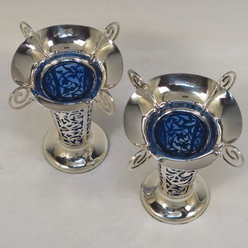 A vert pretty pair of Antique Edwardian Sterling Silver vases, having hand-pierced bodies with floral decoration on round tapering sides, with removable blue-glass liners, with wavy-edge rims and four scroll handles, and all sitting on plain round pedestal feet. These beautiful antique silver vases were made by Harrison and Hipwood of Birmingham in 1910. The dimensions of this fine hand-made pair of antique silver vases are height 13 cms (5.25 inches), and diameter at top 10 cms (4 inches).   