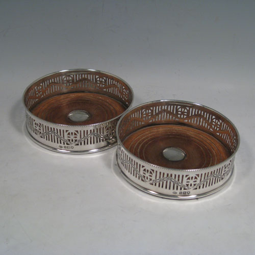 Sterling silver pair of wine bottle table coasters, having round bodies with hand-pierced decoration and engraved swags, with wood-turned bases and silver buttons, and applied beaded borders. Made by Henry Freeman Ltd., of London in 1913. The dimensions of these wine coasters are diameter 13 cms (5 inches), height 4 cms (1.5 inches).   