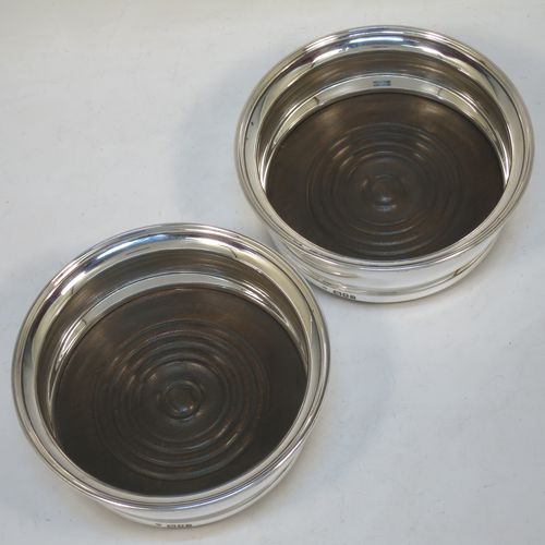 A handsome Sterling Silver pair of wine bottle table coasters, in a plain Georgian style, having round bellied bodies with applied reeded borders and with oak wood-turned bases. Made by C. J. Vander of London in 1960. The dimensions of these fine hand-made silver wine bottle coasters are diameter 14.5 cms (5.75 inches), and height 5 cms (2 inches).    