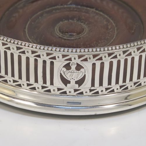 A very pretty Antique Georgian Neoclassical style Sterling Silver wine bottle table coaster, having a round hand-pierced body with geometrical and tureen style decoration, together with an applied bead-edged top border, and an wood-turned base. This beautiful antique silver wine bottle coaster was made in London in 1786. The dimensions of this fine hand-made antique silver wine bottle coaster are diameter 12 cms (4.75 inches), and height 3.5 cms (1.3 inches).   